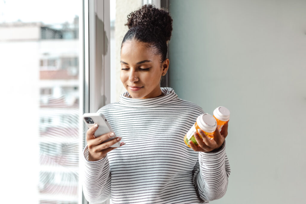 A young woman standing by the window and researching medications on her mobile phone.